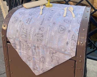 May include: A brown metal trash can with a white and blue patterned tablecloth on top. The tablecloth features a winter holiday theme with snowflakes, penguins, bears, and the words "Happy Holidays" and "Let it Snow".
