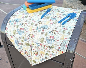 May include: A brown metal trash can with a colorful patterned tablecloth draped over the top. On the tablecloth are three blue, yellow, and orange rectangular containers and two blue plastic utensils.