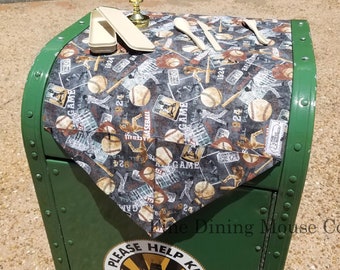 May include: A green metal trash can with a baseball-themed tablecloth on top. The tablecloth has a white candle, a wooden spoon, and a white rectangular box on it. The trash can has a yellow sticker that says "Please Help Keep Our City Clean".