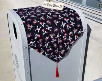 May include: A black and white pirate skull and crossbones patterned table runner with a red tassel. The runner is on top of a gray trash can. A wooden sign reads "Fine Dining Mouse Co."