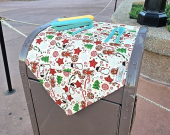 May include: A brown metal trash can with a white and red Christmas themed tablecloth on top. The tablecloth has a pattern of gingerbread men, candy canes, and snowflakes. There are two blue plastic spoons on the tablecloth.