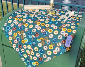 May include: A green tablecloth with a pattern of colorful badges. The tablecloth is on a green mailbox with a white edge. The tablecloth has a white border and the text "The Trading Post" in white letters.