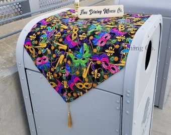 May include: A black table runner with a Mardi Gras theme. It features gold, purple, green, and pink masquerade masks, fleur-de-lis, and beads. The runner has a gold tassel and a sign that reads "Fine Dining Mouse Co."