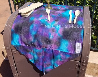 May include: A purple and blue galaxy print table runner with a white candle, a wooden fork, knife, and spoon, and a white container with a lid. The text "FINE DINING MOUSE CO" is printed on the table runner.