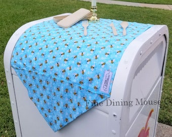May include: A blue and white table runner with a bee pattern. The runner is on a white trash can with a palm tree logo. The runner is decorated with a small wooden box, a candle holder, and wooden utensils.