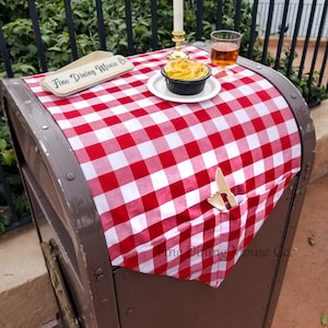 May include: A red and white checkered tablecloth covers a brown metal trash can. A small plate with a golden brown pastry and a lit candle are on the tablecloth. A glass of amber liquid sits next to the plate. A sign reads "Fine Dining Mouse Co."