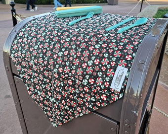 May include: A black and green fabric placemat with a peppermint candy design. The placemat is folded and placed on top of a brown metal trash can. A blue plastic cutlery set is on top of the placemat.