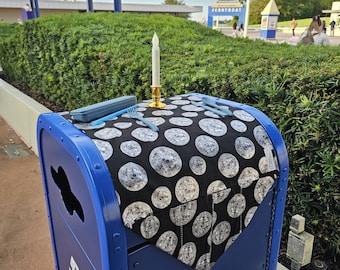 May include: A blue recycling bin with a black and white patterned tablecloth. A lit candle in a gold holder sits on the tablecloth, along with blue utensils and a blue container. The background includes green bushes and a building.