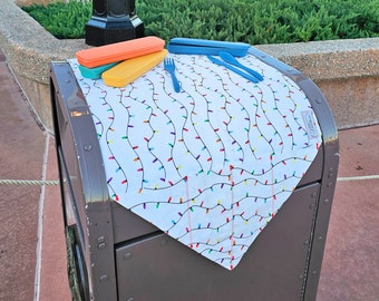 May include: A white tablecloth with a colorful string of lights pattern, a blue and a yellow plastic utensil set, and three orange and yellow plastic containers on top of a brown metal trash can.