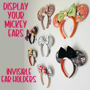May include: A white wall with the text "DISPLAY YOUR MICKEY EARS INVISIBLE EAR HOLDERS" in pink letters.  Seven pairs of Mickey Mouse ears are displayed on the wall using clear plastic ear holders. The ears are in various colors and patterns, including black, white, red, green, brown, and gold.