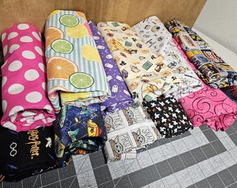 May include: A collection of folded fabric bolts featuring various patterns including pink with white polka dots, citrus fruit slices, purple with ghosts, Disney characters, Star Wars themes, Harry Potter motifs, and space-themed fish prints.