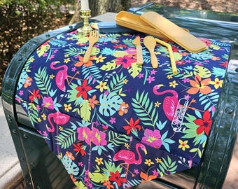 May include: A tropical-themed placemat with pink flamingos, green leaves, and colorful flowers. A set of gold utensils and a straw are laid out on the placemat. A small gold candle holder with a white candle is in the background.