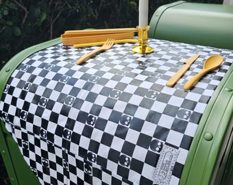 May include: A black and white checkered table runner with a skull pattern. The runner is on a green surface and has a gold candle holder with a white candle in it. There are also wooden utensils on the table runner.