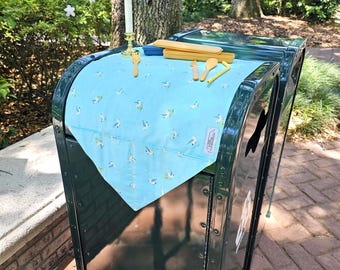 May include: A dark green mailbox with a light blue bird-patterned cloth draped over it. Wooden utensils and a candle holder with a white candle are on top. The scene is outdoors with trees and greenery in the background.