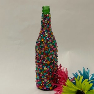 May include: A glass bottle covered in colorful rhinestones, with a green top, and three artificial flowers in pink, blue, and green.