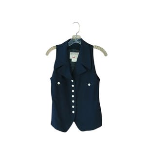 May include: A sleeveless navy blue vest featuring a pointed collar, a row of seven white buttons down the front, and two small chest pockets with white button closures. The interior label displays the text MONDI with a small graphic icon.