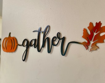 May include: Metal wall decor with the word "gather" in black script, a small orange pumpkin on the left, and a large orange leaf on the right.