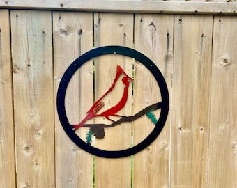May include: A red cardinal bird metal silhouette on a black circular background with a branch and pine needles. The metal art is attached to a wooden fence.