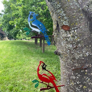 May include: Two metal bird silhouettes mounted on a tree. The blue jay is perched on a branch with green leaves. The cardinal is perched on a branch with green leaves. Both birds are facing to the right.