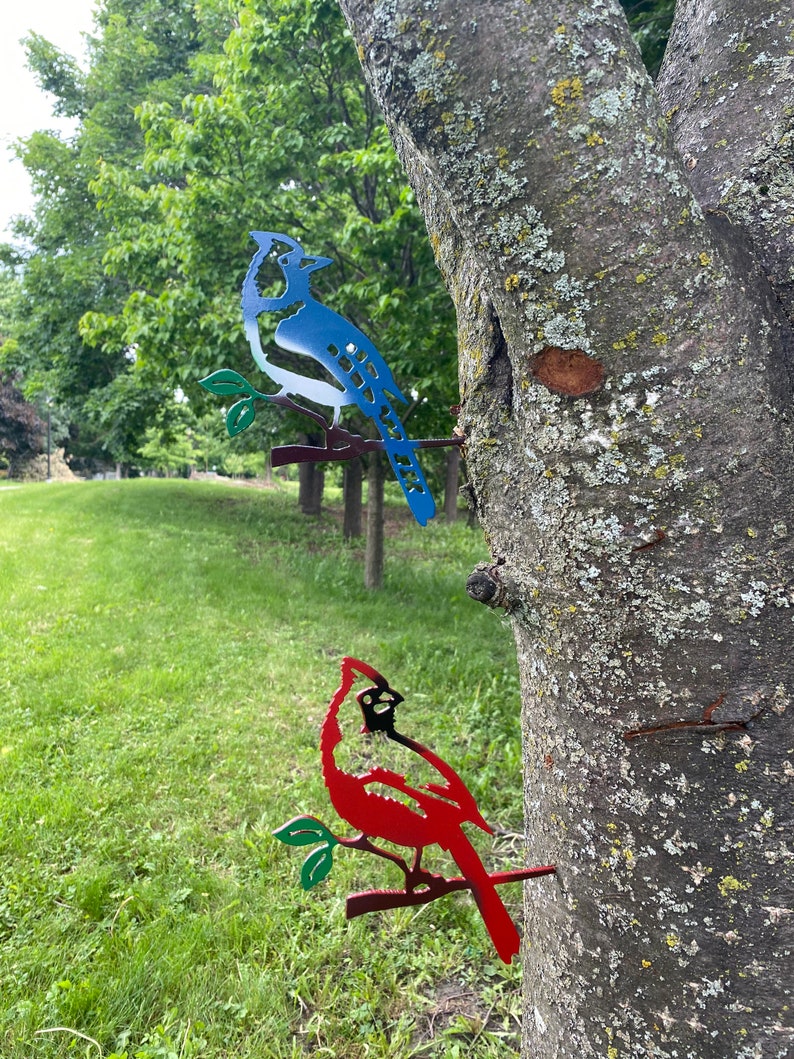 May include: Two metal bird silhouettes mounted on a tree. The blue jay is perched on a branch with green leaves. The cardinal is perched on a branch with green leaves. Both birds are facing to the right.
