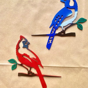 May include: Two metal bird silhouettes on a brown background. The blue jay is perched on a branch with green leaves. The cardinal is perched on a branch with green leaves. Both birds are painted in their respective colors.