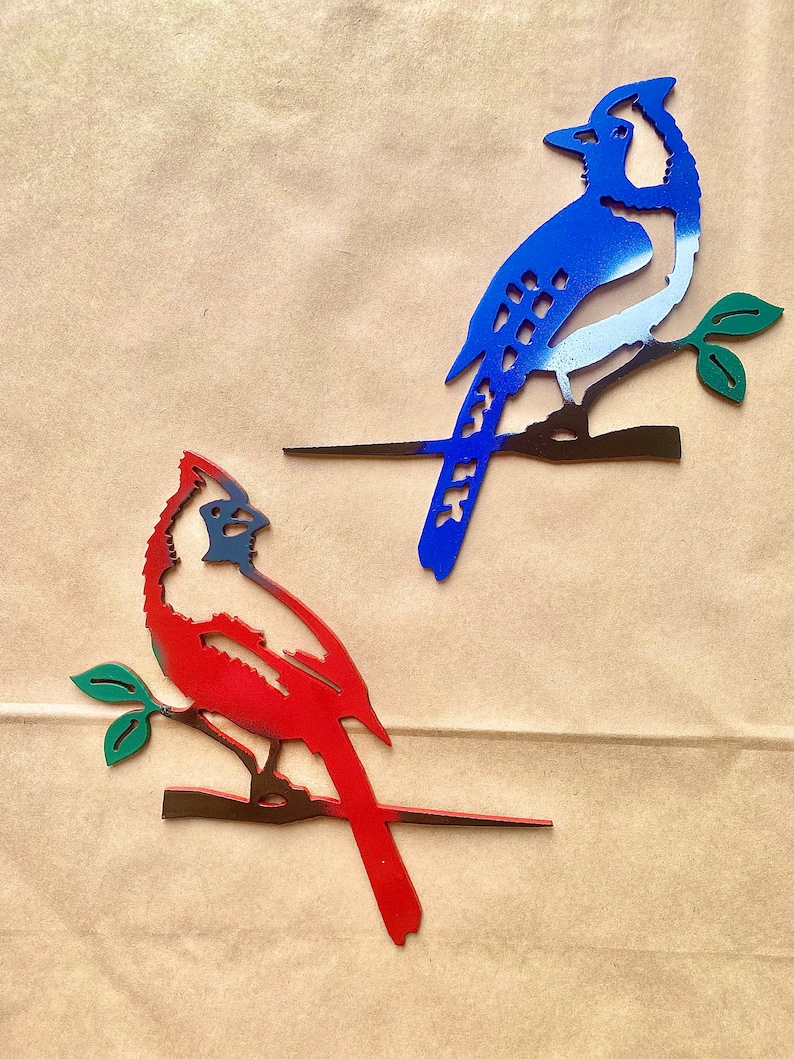 May include: Two metal bird silhouettes on a brown background. The blue jay is perched on a branch with green leaves. The cardinal is perched on a branch with green leaves. Both birds are painted in their respective colors.