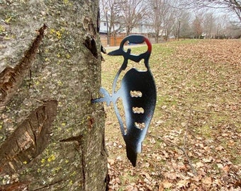 May include: A metal woodpecker silhouette is attached to a tree trunk. The bird is black and white with a red head. The tree bark is brown and textured, with green and yellow patches of lichen. The background shows a park with bare trees and fallen leaves.