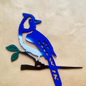 May include: A blue jay metal art piece. The bird is perched on a branch with green leaves. The bird's body is blue, with white and black accents. The metal art is designed to be mounted on a wall or other surface.