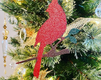 May include: Red glitter cardinal bird Christmas ornament hanging on a green Christmas tree. The cardinal is perched on a brown branch with green leaves.