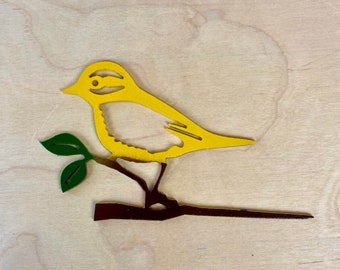 May include: A yellow bird silhouette with a dark brown branch and green leaves. The bird is perched on the branch, and the background is a light wood grain. The bird's eye is visible.