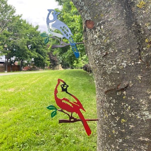 May include: Two metal bird silhouettes mounted on a tree trunk. A blue jay with a white chest is perched on a branch on the top. A red cardinal is perched on a branch below the blue jay.