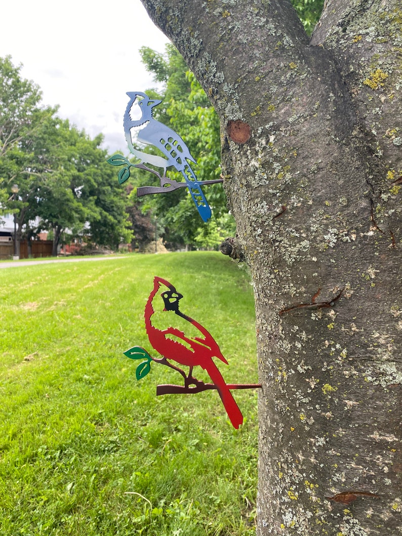 May include: Two metal bird silhouettes mounted on a tree trunk. A blue jay with a white chest is perched on a branch on the top. A red cardinal is perched on a branch below the blue jay.