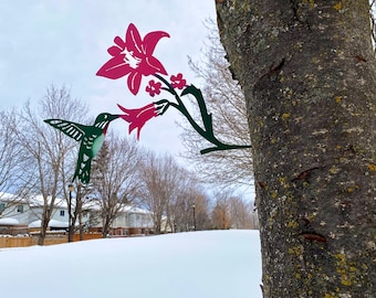 May include: A metal hummingbird sculpture with outstretched wings, perched on a tree branch, is about to feed from a pink metal flower. The sculpture is attached to a tree trunk with a brown bark and some green moss.