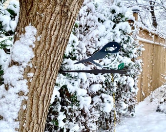 May include: A black metal bird silhouette with a green leaf attached to a branch, mounted on a tree trunk covered in snow. The background is a snowy winter scene.