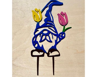 May include: A blue metal gnome with a yellow and pink tulip in each hand. The gnome is standing on two black metal stakes.