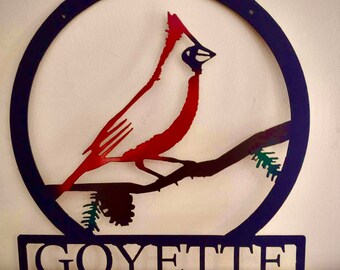 May include: Metal wall art featuring a red cardinal perched on a branch with pine needles. The cardinal is inside a black circle with a black nameplate below that reads "GOYETTE".