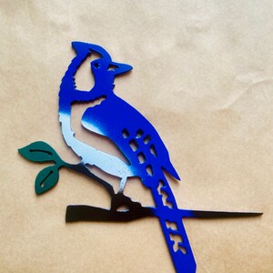 May include: Blue jay metal silhouette with white and black accents, perched on a branch with green leaves. The bird is painted in a gradient style, with blue fading to white on the body.