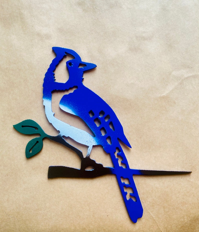 May include: Blue jay metal silhouette with white and black accents, perched on a branch with green leaves. The bird is painted in a gradient style, with blue fading to white on the body.