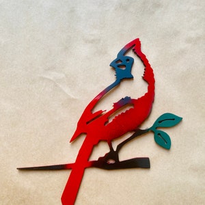 May include: A red and black metal cardinal silhouette perched on a branch with green leaves. The cardinal is facing to the right and has a long tail.