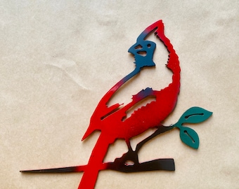 May include: A red and black metal cardinal silhouette perched on a branch with green leaves. The cardinal is facing to the right and has a long tail.