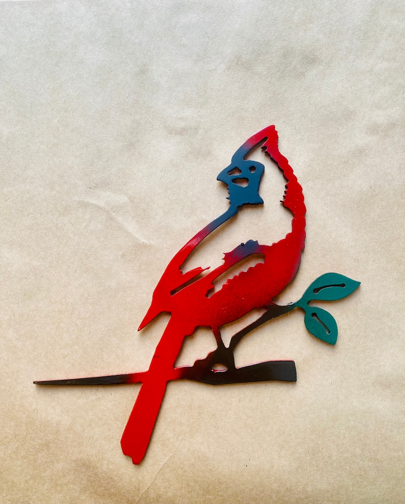 May include: A red and black metal cardinal silhouette perched on a branch with green leaves. The cardinal is facing to the right and has a long tail.
