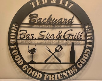 May include: Black metal sign with the text "TED & LIZ Backyard Bar. Spa & Grill GOOD FOOD GOOD FRIENDS GOOD TIMES" in a circular design. The sign features a wine glass, a spatula and a fork, and a bottle.