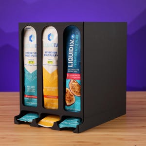 Peut inclure: Un présentoir noir contient trois sachets de Liquid I.V. Hydration Multiplier en bleu, jaune et turquoise. Les sachets sont présentés dans un présentoir noir avec des fentes individuelles et un petit plateau en bas pour chaque saveur. Le fond est un dégradé de violet.