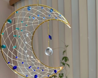 May include: A crescent moon-shaped dream catcher with a gold-colored frame. The web is white, adorned with blue, teal, and clear beads. A clear crystal ball and a blue crystal hang from the center. The dream catcher is suspended by a gold chain.