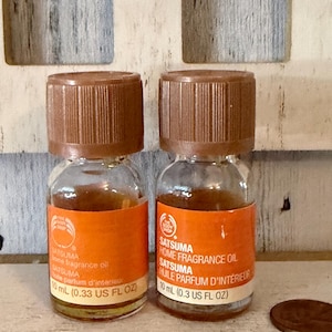 May include: Two small glass bottles of The Body Shop Satsuma home fragrance oil, each featuring a brown screw-top cap and an orange label with white text stating 10 mL (0.33 US FL OZ). The bottles contain a golden-colored liquid fragrance oil.