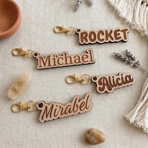 May include: Wooden keyrings with the names "Rocket", "Michael", "Alicia", and "Mirabel" in a decorative font. Each keyring has a gold-coloured clasp. The keyrings are made of light-coloured wood with darker wood for the lettering.