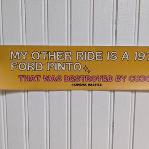 May include: A yellow bumper sticker with pink text that reads "My other ride is a 1978 Ford Pinto that was destroyed by Cujo" with the social media handle "@cinema_nasties" at the bottom.