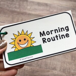 Pu&ograve; includere: Una carta bianca con un'illustrazione di un sole verde e giallo e il testo "Morning Routine" in nero. La carta ha un anello verde attaccato.