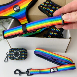 May include: A rainbow dog harness, leash, and poop bag holder with the text "House of Dawg" on each item. The harness and leash are both made of a rainbow striped fabric with black paw prints on the harness. The poop bag holder is black with rainbow paw prints.