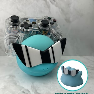 May include: A clear plastic video game controller with a blue and white character figurine attached to the front. The figurine has a pair of sunglasses with black and white stripes. The text "FREE DITTO SQUAD WITH PURCHASE" is visible below the figurine.